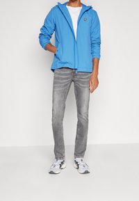 Light blue zip-up hooded jacket with pockets, worn over a white t-shirt, paired with gray jeans and multicolored athletic shoes.