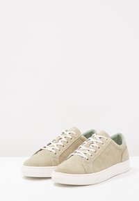 Světlé béžové semišové tenisky s bílými gumovými podrážkami, se béžovými tkaničkami, jemnými akcenty vzadu a značkovým detailem na boku. Hladký povrch.