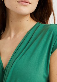 Zelený top s krátkým rukávem a výstřihem do V. Vyrobeno z měkké, lehké tkaniny s hladkým povrchem. Minimální detaily a přiléhavý střih.
