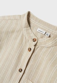 Camicia button-up beige a righe con colletto rotondo, dotata di due bottoni marroni e una tasca sul petto. Realizzata in tessuto testurizzato.
