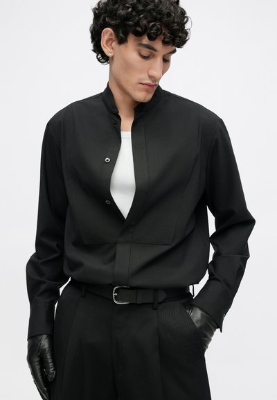 Μαύρο πουκάμισο με μακρύ μανίκι, ψηλό γιακά και κλείσιμο με κουμπιά, με σχεδιασμό επίπεδης τσέπης και λεία υφή, συνδυασμένο με μαύρη ζώνη.