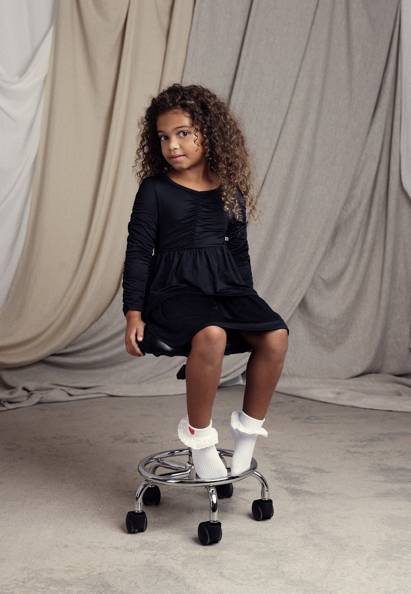 Μαύρο μακρύ φόρεμα με στυλ μπροστά, φαρδύ φούστα, φορεμένο με λευκές κάλτσες με φουσκωτό σχέδιο που διαθέτουν μια κόκκινη καρδιά, καθισμένο σε ένα χρωμιωμένο σκαμπό.