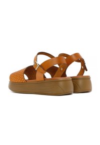 Sandali in pelle marrone chiaro con texture intrecciata, lati aperti e cinturini regolabili alla caviglia. Presentano una suola piatta spessa, di colore marrone, con righe orizzontali.