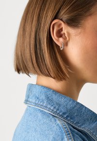 Cercel din argint cu o suprafață texturată, purtat pe părul scurt și drept de culoare brunete. Subiectul poartă o jachetă din denim de culoare bleu deschis.