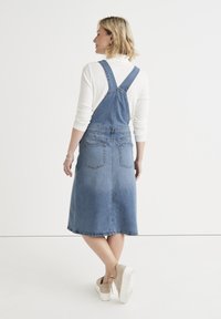 Sininen farkkuhaalari, polvipituinen, säädettävillä olkahihnoilla, kahdella takataskulla ja levenevällä helmalla. Yhdistetty vaaleanbeigeihin korokepolkimiin.