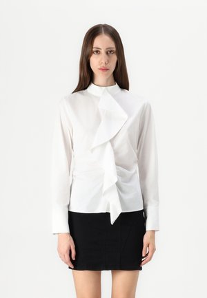 Hvit bluse med lange ermer og høy krage, med en fremtredende volang foran. Modellen har parret den med et svart tettsittende skjørt.