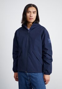 Veste à capuche marine en tissu léger, dotée d'un zip sur le devant, de poignets élastiques et de deux poches avant angulaires. Logo sur la manche.