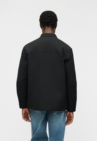 Άνδρας με μαύρο μπουφάν και μπλε τζιν, που στέκεται με την πλάτη στραμμένη προς τον θεατή σε λευκό φόντο.