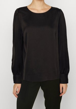 Blouse noire à manches longues avec épaules légèrement bouffantes et larges poignets, portée rentrée dans un pantalon noir par une personne debout devant un fond uni.