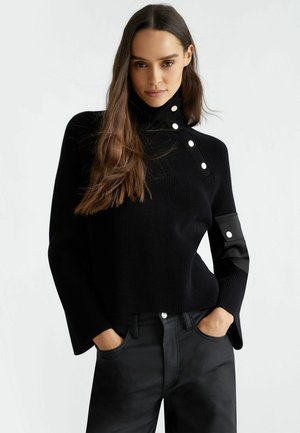 Maglione nero a maglia con colletto alto e dettagli in bottone, caratterizzato da maniche ampie e una finitura testurizzata. Abbinato a pantaloni neri.