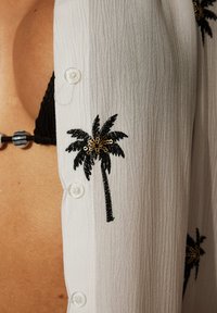 Camisa blanca ligera con tejido texturizado, que presenta bordados de palmeras en negro y dorado. Botones transparentes en la parte delantera.
