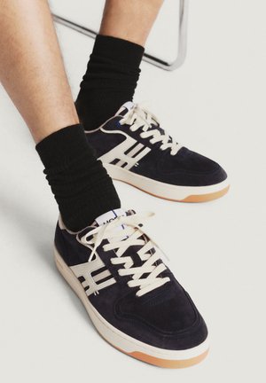 Sneakers in suede blu navy con strisce bianche, dotati di lacci crema e suole in gomma. Indossati con calzini neri a coste alla caviglia. Cuciture dettagliate ben visibili.