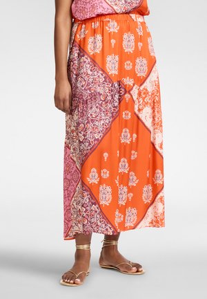 Vrouw die een maxi rok draagt met oranje, roze en paarse patchworkpatronen en gouden platte sandalen met bandjes op een witte achtergrond.