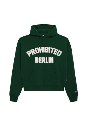 Mand iført en mørkegrøn hættetrøje med "PROHIBITED BERLIN" tekst, grå bukser og en hvid strikket hue med en mærkat, står med hænderne i lommerne.