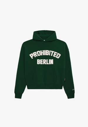 Mand iført en mørkegrøn hættetrøje med "PROHIBITED BERLIN" tekst, grå bukser og en hvid strikket hue med en mærkat, står med hænderne i lommerne.