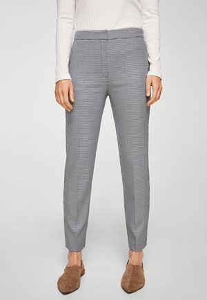 Femme portant un pantalon slim à carreaux noirs et blancs, longueur cheville, avec un haut blanc à manches longues côtelé et des chaussures marron sans lacets.