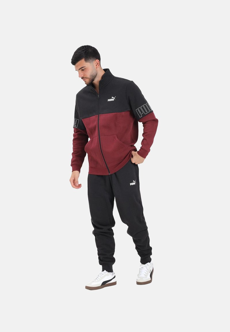Uomo che indossa una giacca con zip Puma nera e bordeaux, pantaloni della tuta Puma neri e sneakers bianche, in piedi su uno sfondo bianco uniforme.