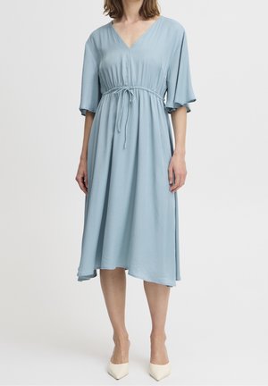 Nainen pukeutuneena vaaleansiniseen midi-mekkoon, jossa on V-kaula-aukko, puolihihaiset hihat ja kiristysnyörivyötärö, yhdistettynä valkoisiin teräväkärkisiin korkokenkiin.