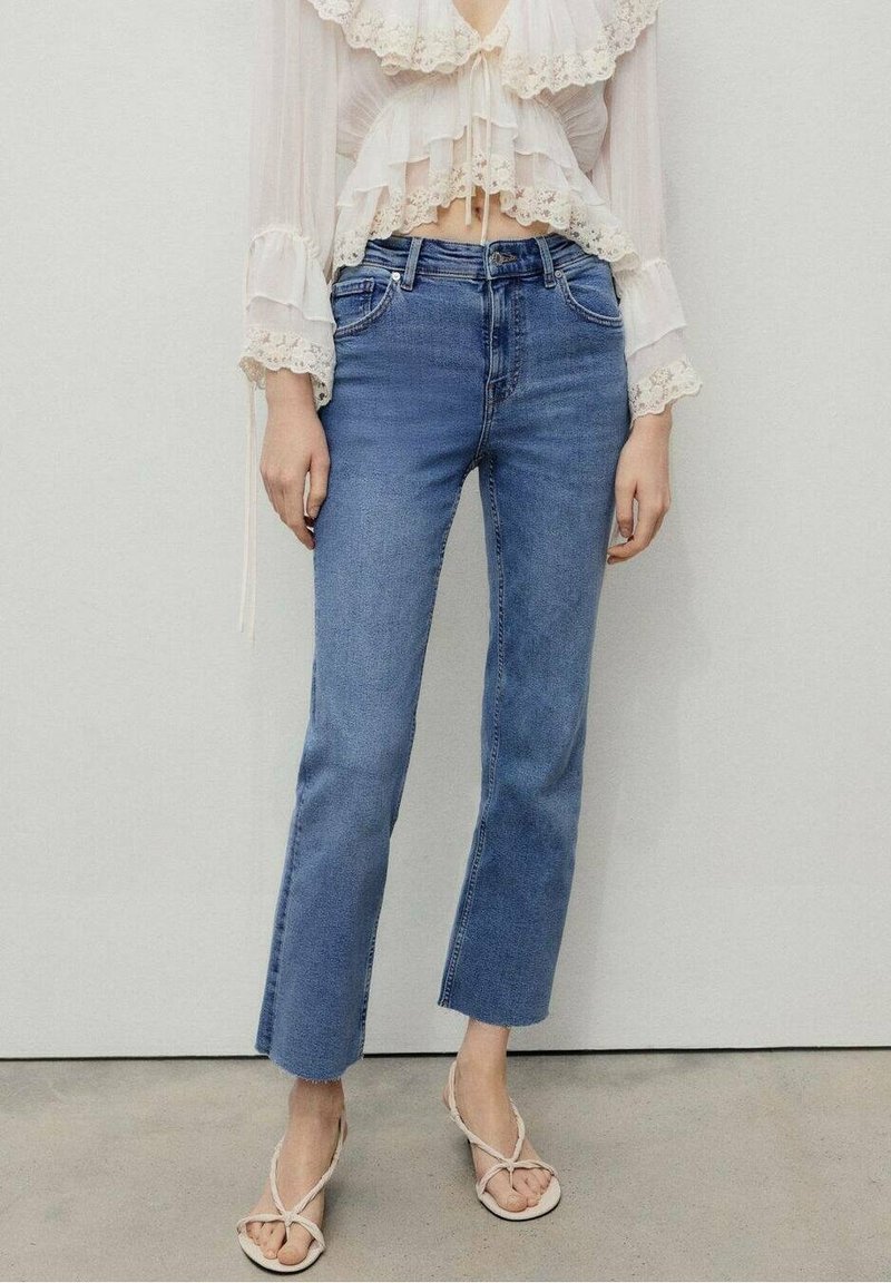 Hellblaue Jeans im geraden Schnitt mit ausgefranstem Saum, kombiniert mit einer transparenten, rüschierten weißen Bluse und hellen Sandalen.