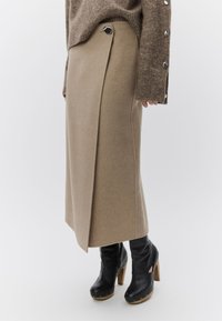 Μπεζ midi φούστα τύπου wrap με μαύρη λεπτομέρεια κουμπιού, συνδυασμένη με καφέ ζακέτα και μαύρα δερμάτινα μποτάκια με ψηλό τακούνι, σε λευκό φόντο.