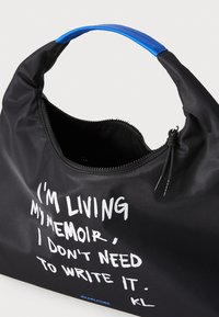 Black nylon bag with a blue handle, featuring white text: "I'M LIVING MY MEMOIR, I DON'T NEED TO WRITE IT." Zippered closure, smooth texture.