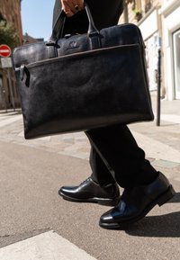 Maletín de cuero negro con una superficie texturizada, doble asa en la parte superior y un cierre de cremallera plateado. Llevado junto a zapatos negros pulidos y pantalones a medida.