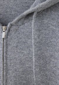 Gros plan d’un tissu tricoté gris avec une fermeture éclair métallique et un cordon, montrant la texture et les détails des coutures.