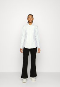 Camisa blanca de manga larga con botones y cuello, combinada con pantalones negros acampanados y zapatillas blancas. Tejido suave, corte ajustado.