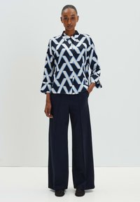 Camicia a fantasia in navy e bianco. Tessuto simile alla seta, con linee geometriche blu. Abbinata a pantaloni a gamba larga navy e scarpe nere.
