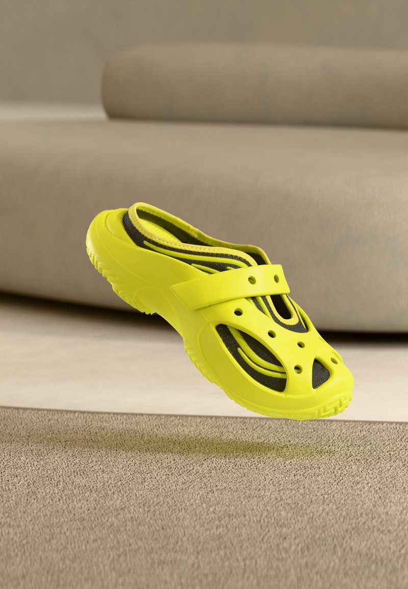 Φωτεινό κίτρινο σανδάλι τύπου slip-on με οπές αερισμού, μαύρη εσωτερική σόλα, αιωρείται πάνω από μπεζ χαλί μπροστά από μια μπεζ καναπέ.