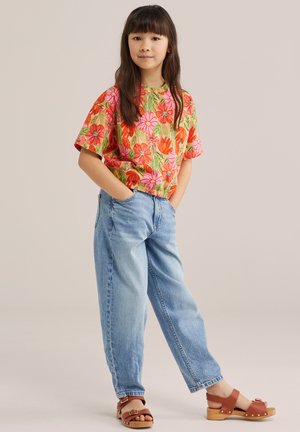 Fată în picioare, cu mâinile în buzunare, purtând un top roșu cu flori, blugi bleu deschis și sandale cu talpă din lemn maro, pe un fundal simplu.