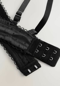 Czarny stanik wykończony koronką, z miękkiego materiału, z regulowanymi ramiączkami, w wzór w kropki oraz z zapięciem na haftki z metalowymi akcentami.