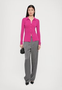 Fuksia cardigan med knapper, sammen med svarte og hvite mønstrede vide bukser. Modellen holder en liten svart håndveske.