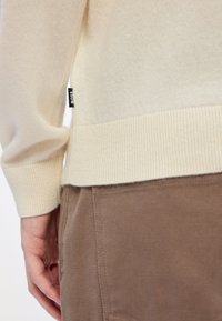 Pulover crem cu tiv și manșete cu nervuri, realizat dintr-un material moale. Prezintă o mică etichetă neagră „BOSS” pe lateral. Pantaloni maro afișați.