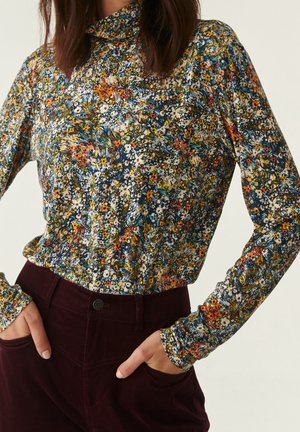 Kvinde iført bluse med lange ærmer og høj hals med flerfarvede små blomster, kombineret med mørkelilla bukser med høj talje, hænderne i lommerne.