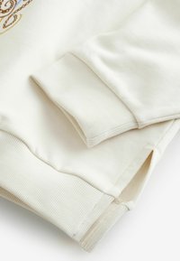 Krémovou farbou mikinovina s rebrovanými manžetami a bočným rozparkom. Hladká textúra so spevneným šitím na leme.