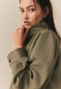 Oliwkowa koszula z kołnierzykiem, z guzikami przy mankietach i miękką teksturą. Posiada marszczone ramiona i luźny fason. Model trzyma kołnierzyk.