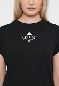 Tricou negru cu mânecă scurtă, din bumbac, având un grafic alb care spune "REPLAY NOT ORDINARY" cu un logo deasupra.
