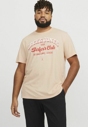 Lys ferskenfarvet T-shirt med korte ærmer, prydet med fed rød grafisk tekst "JACK & JONES Surfer's Club" og logo på fronten, parret med sorte bukser.