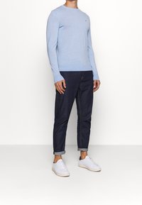 Homme portant un pull léger bleu à manches longues, un jean foncé retroussé et des baskets blanches, se tenant devant un fond blanc uni.