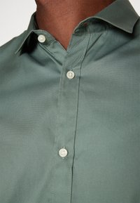 Chemise verte à col boutonné, fabriquée en tissu lisse, avec un col classique et des boutons en plastique blanc, ornée de détails de couture subtils.
