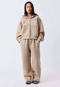 Sudadera beige con cremallera, grandes bolsillos delanteros y pantalones a juego de corte holgado. Tejido suave, diseño oversize, acento de marca minimalista.