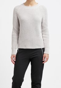 Eine Person trägt einen hellgrauen, gerippten Strickpullover und schwarze Hosen mit Reißverschlussdetails an den Taschen und steht vor einem weißen Hintergrund.