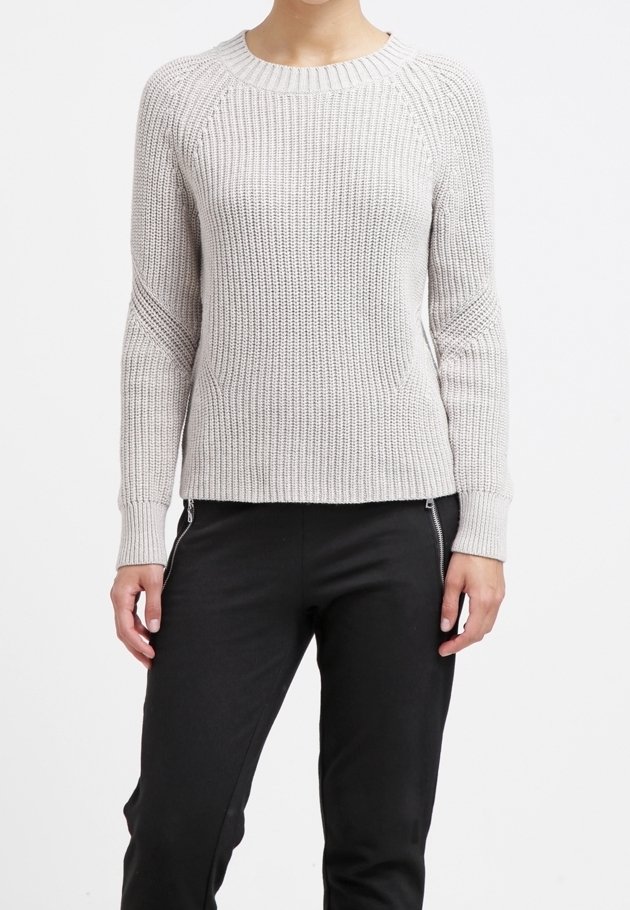 Eine Person trägt einen hellgrauen, gerippten Strickpullover und schwarze Hosen mit Reißverschlussdetails an den Taschen und steht vor einem weißen Hintergrund.