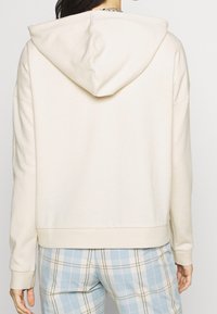Sweat à capuche beige clair avec une capuche à cordon et des poignets côtelés. Tissu doux et texturé, présentant une coupe décontractée et un design court.