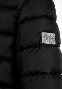 Veste matelassée noire avec un design en piqué, dotée d'un tissu brillant et texturé ainsi qu'une pièce brillante ornée de paillettes multicolores et du texte "Minoti".