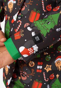 Melnas auduma jakas ar košām Ziemassvētku tēmatiskām zīmēm, kurās redzami ziemeļbrieži, dāvanas un egles. Zaļas aproces akcenti ar logo tagu un pogām.