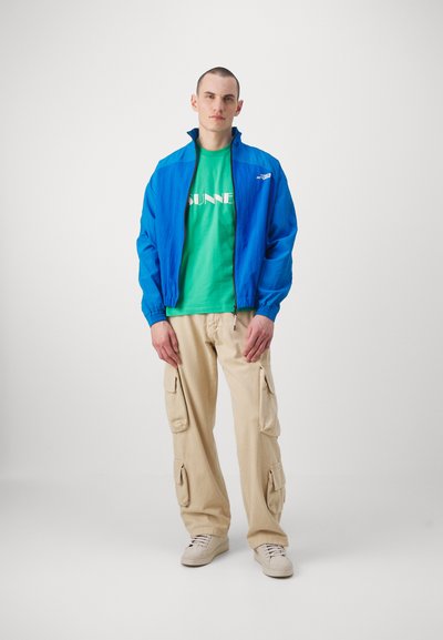 Blå lett zip-up jakke, grønn T-skjorte med hvit tekst, beige cargo-bukser med lommer og beige joggesko. Enkelt design, avslappet passform.