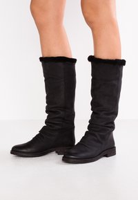 Bottes noires en cuir montantes avec un design froissé, dotées d'une douce bordure en fourrure noire en haut et d'une semelle texturée pour une meilleure adhérence.