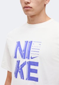 Νέος/νέα που φοράει λευκό T-shirt Nike με μεγάλο pixelated λιλά κείμενο "NIKE" και μικρότερο κείμενο που γράφει "Σχεδιασμένο ακριβώς σύμφωνα με τις προδιαγραφές των πρωταθλητών αθλητών."